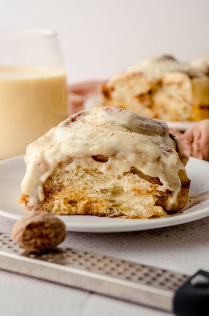 An eggnog cinnamon roll on a plate. There is a nutmeg and a grater in the foreground.