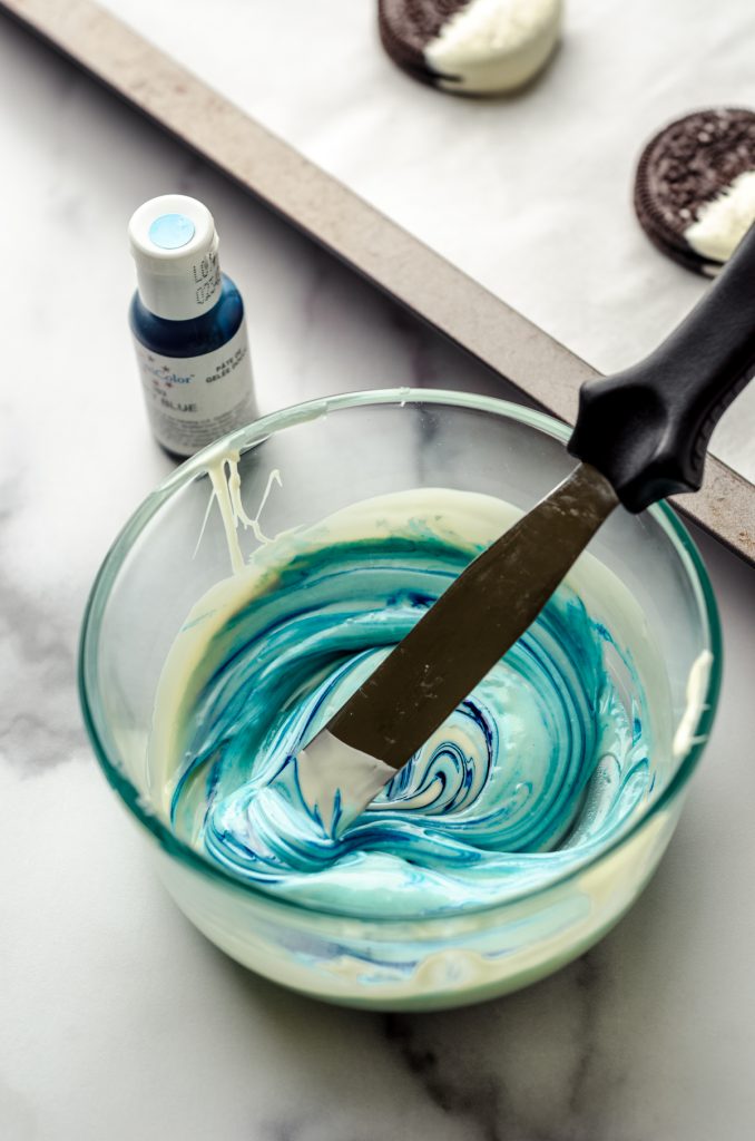 Someone is stirring blue food coloring into a bowl of melted white chocolate to make light blue chocolate.