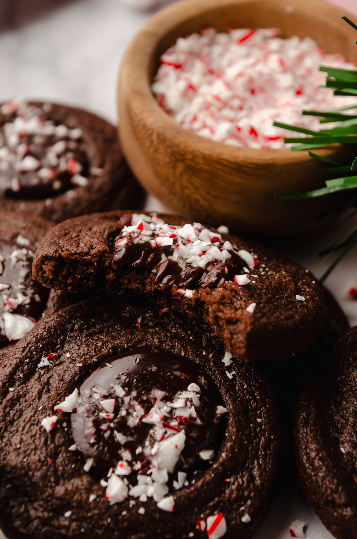 A chocolate peppermint thumbprint cookie that has a bite taken out of it on a surface with a bowl of crushed candy canes in the background.