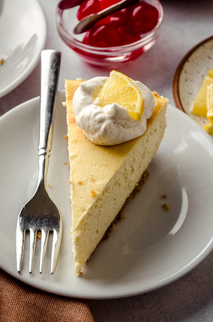 A slice of cheesecake on a plate with a dollop of whipped cream and a slice of lemon on top.