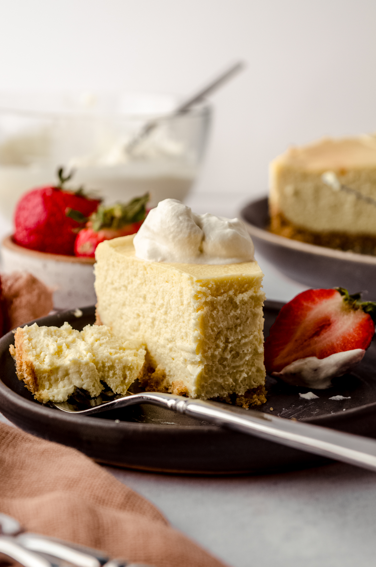 A slice of classic cheesecake with whipped cream on a plate. Someone has taken a bite from the cheesecake with a fork.