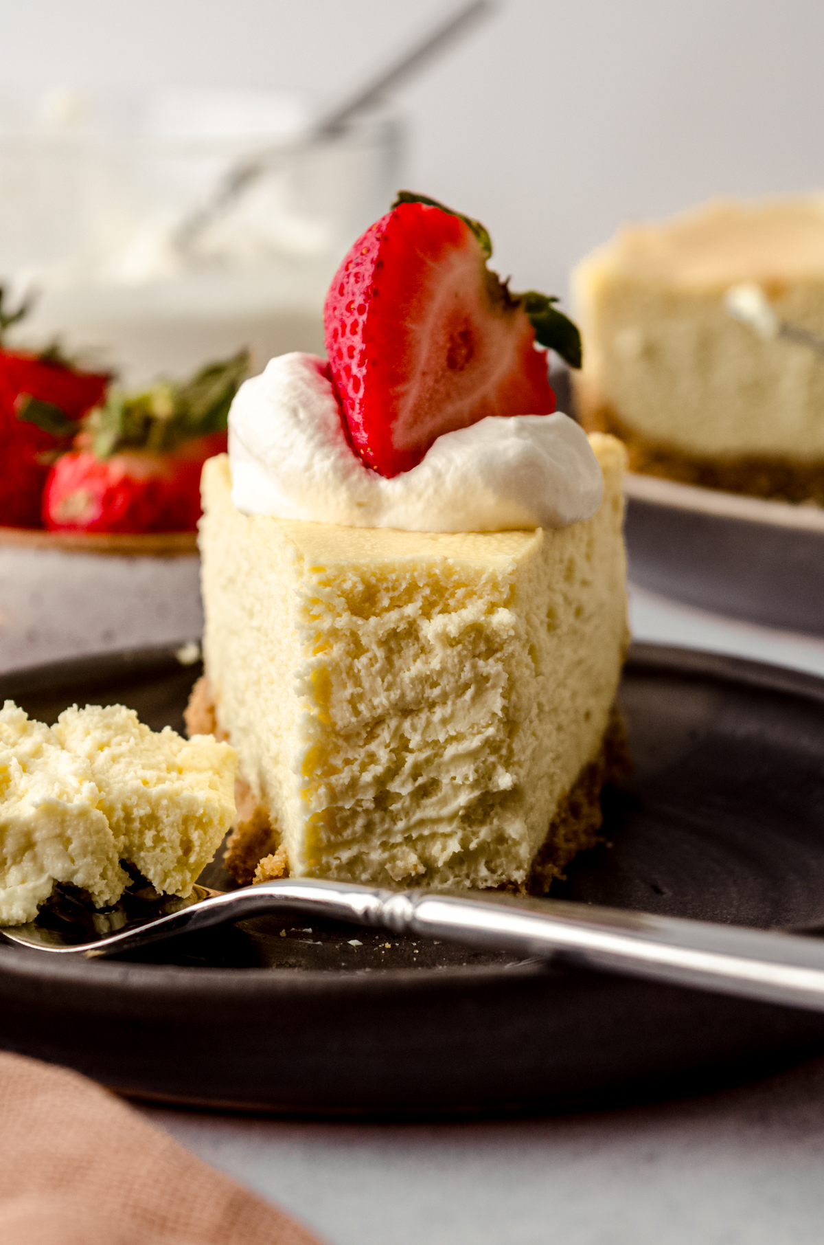 A slice of classic cheesecake with whipped cream and a strawberry on a plate. Someone has taken a bite from the cheesecake with a fork.
