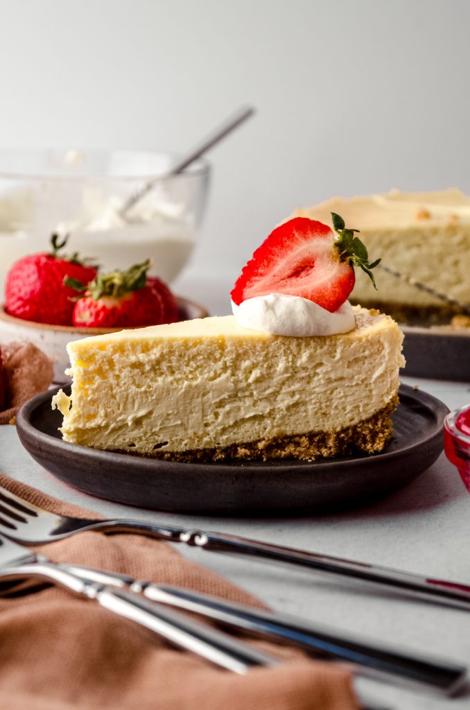 A slice of classic cheesecake on a plate. There is whipped cream and a sliced strawberry on top.