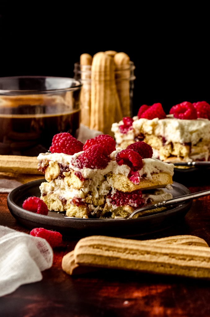 A slice of raspberry tiramisu on a plate with a fork that has a bite taken out of it. There is a cup of coffee and a jar of ladyfingers in the background.