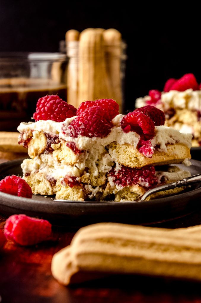A slice of raspberry tiramisu on a plate with a fork that has a bite taken out of it. There is a cup of coffee and a jar of ladyfingers in the background.