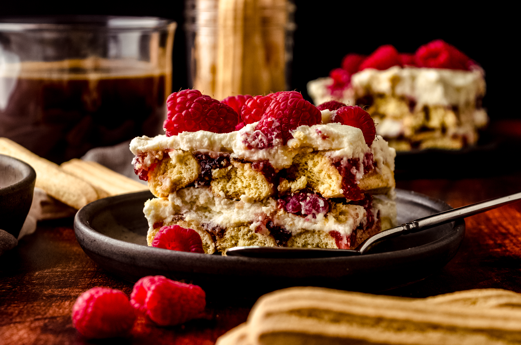 A slice of raspberry tiramisu on a plate with a fork. There is a cup of coffee and a jar of ladyfingers in the background.
