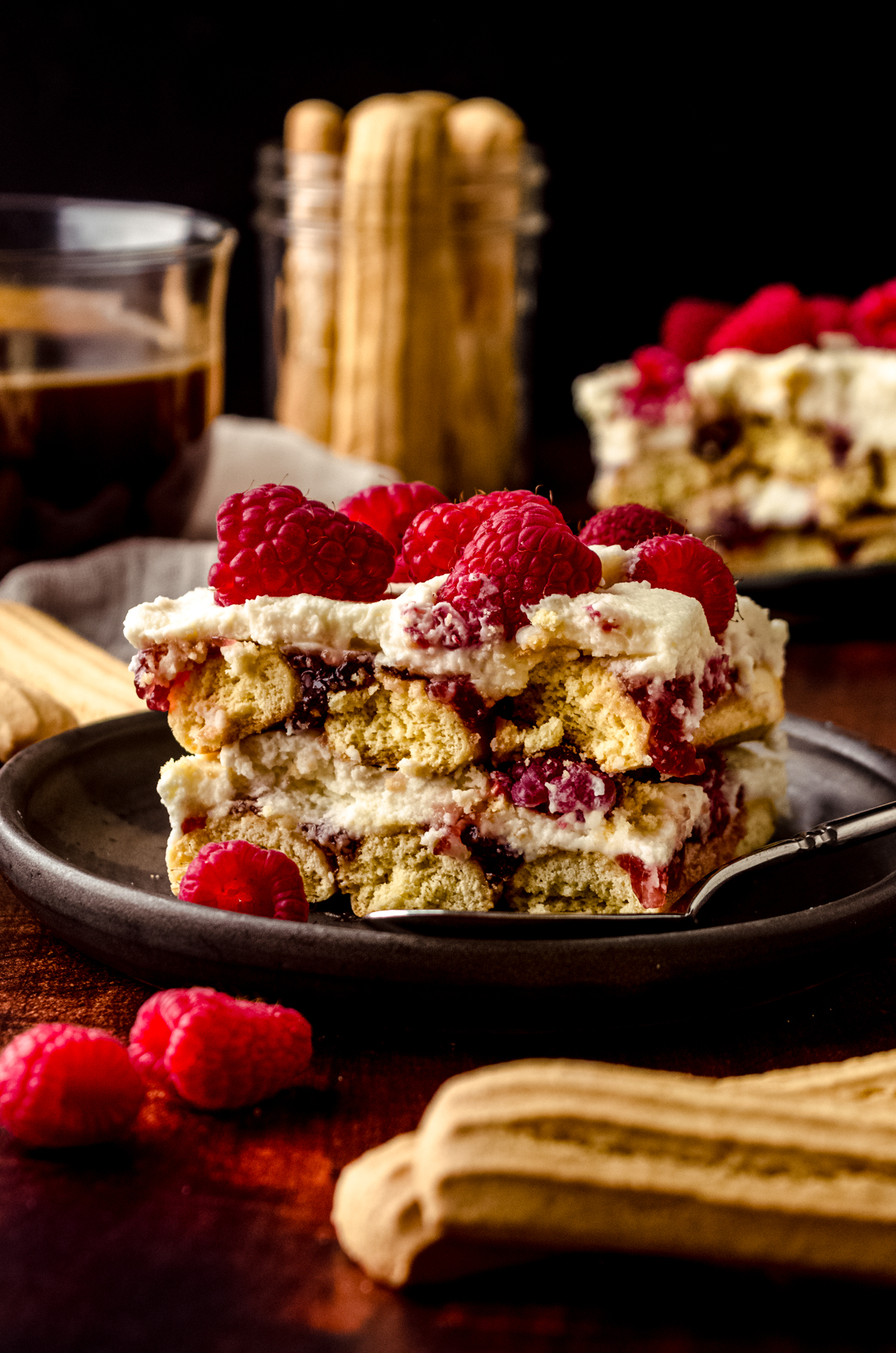 A slice of raspberry tiramisu on a plate with a fork. There is a cup of coffee and a jar of ladyfingers in the background.
