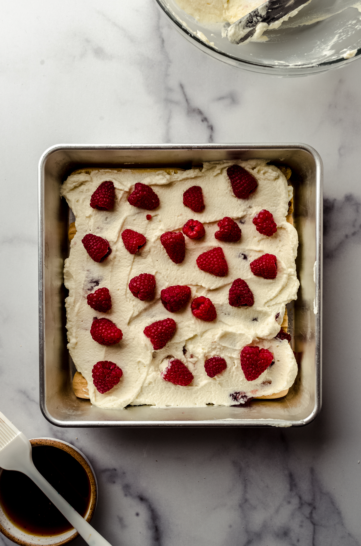 Aerial photo of a layer of mascarpone cream and raspberries on top of a layer of ladyfingers and raspberry preserves in the bottom of a square baking pan as the base for raspberry tiramisu.