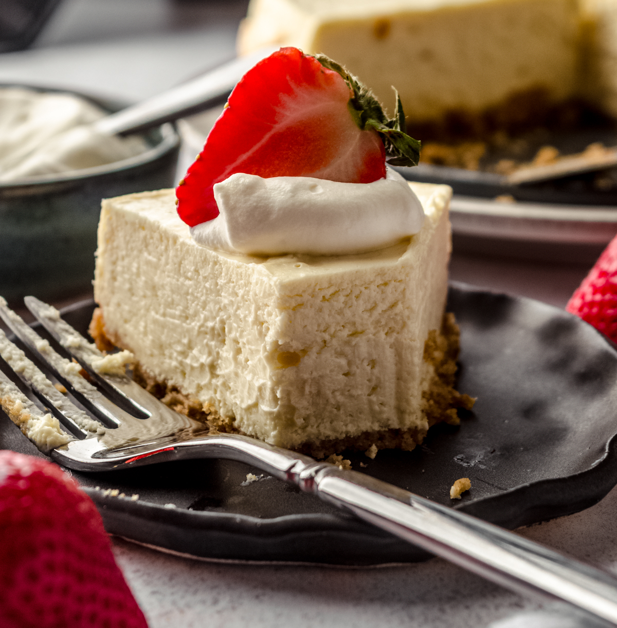 A slice of Instant Pot cheesecake on a plate with a fork. There is a dollop of whipped cream and a strawberry on top. A bite has been taken out of it.