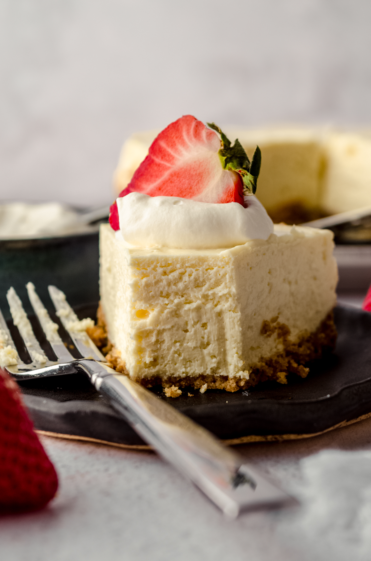 A slice of Instant Pot cheesecake on a plate with a fork. There is a dollop of whipped cream and a strawberry on top. A bite has been taken out of it.