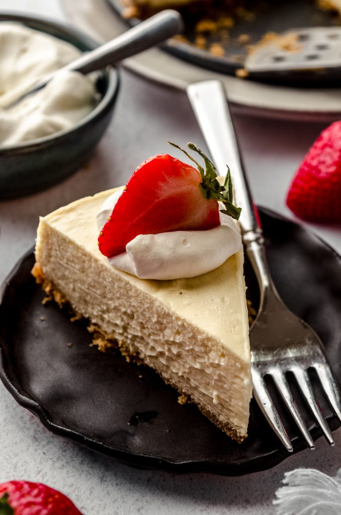 A slice of Instant Pot cheesecake on a plate with a fork. There is a dollop of whipped cream and a strawberry on top.