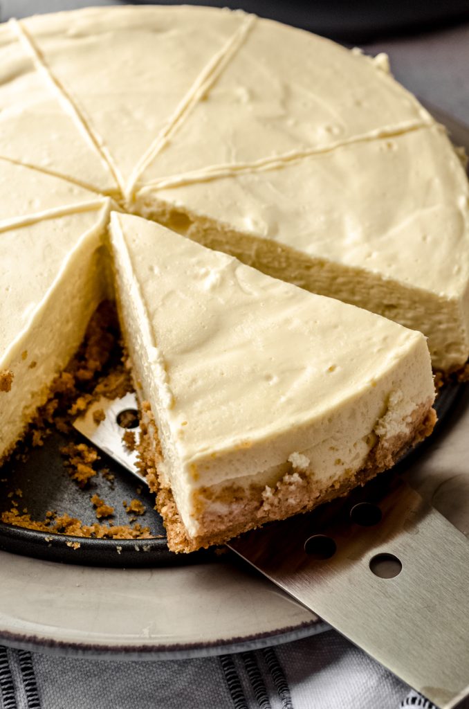 An Instant Pot cheesecake on a plate. It has been sliced and someone is using a spatula to take a piece out.