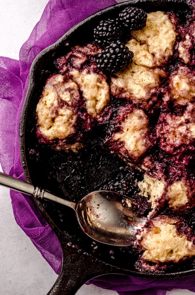 Someone has used a spoon to take a portion of blackberry dumplings out of a cast iron skillet.