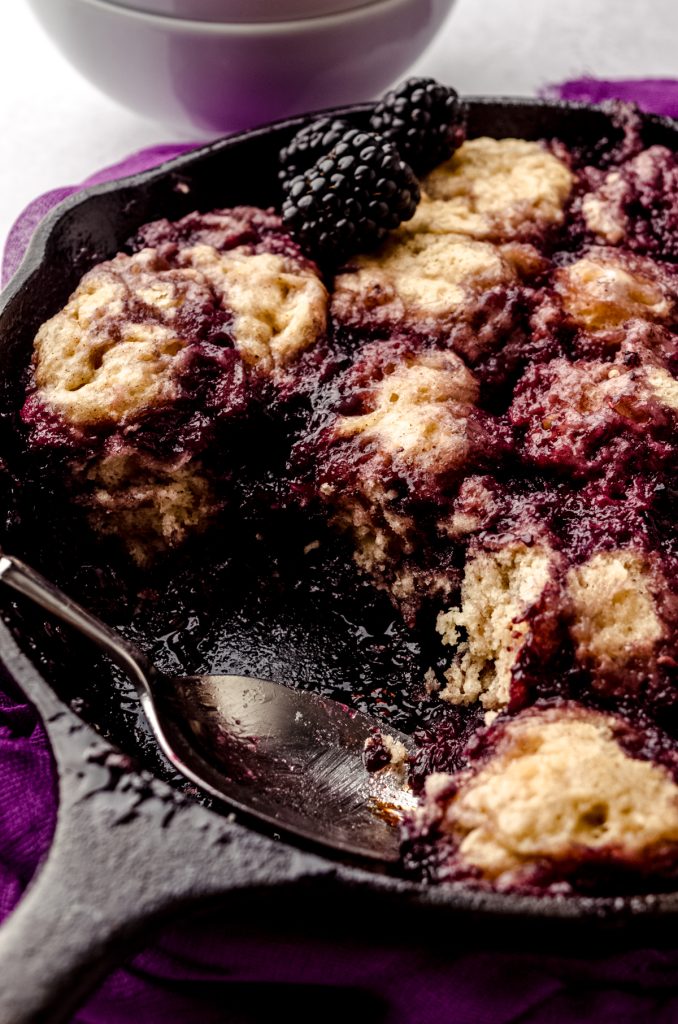 Someone has used a spoon to take a portion of blackberry dumplings out of a cast iron skillet.