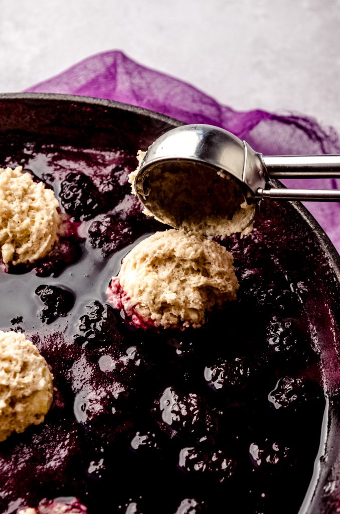 Someone is using a small cookie scoop to drop balls of dough onto cooked blackberry mixture to make blackberry dumplings.