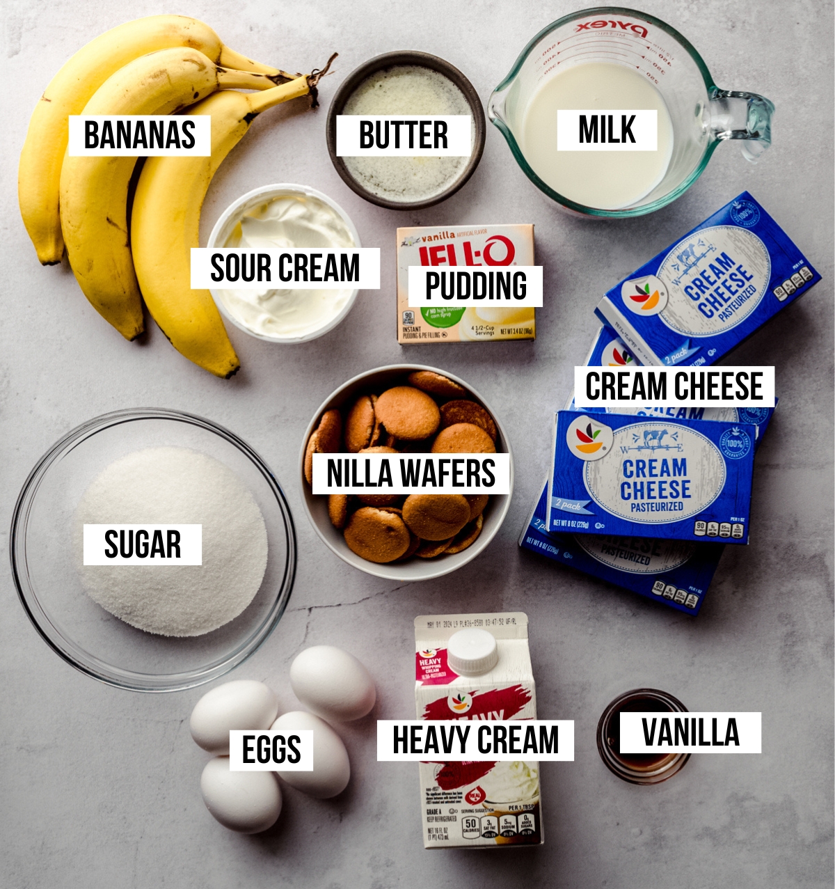 Aerial photo of ingredients to make banana pudding cheesecake on a white surface with text overlay labeling each ingredient.