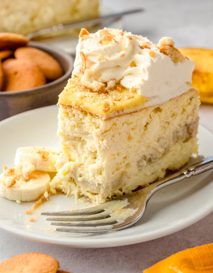 A slice of banana pudding cheesecake on a plate with a fork and a bite has been taken out of the slice.