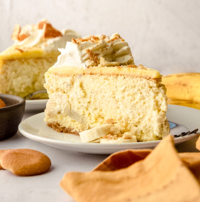 A slice of banana pudding cheesecake on a plate with another piece in the background and Nilla Wafers and an orange-yellow kitchen towel in the foreground.