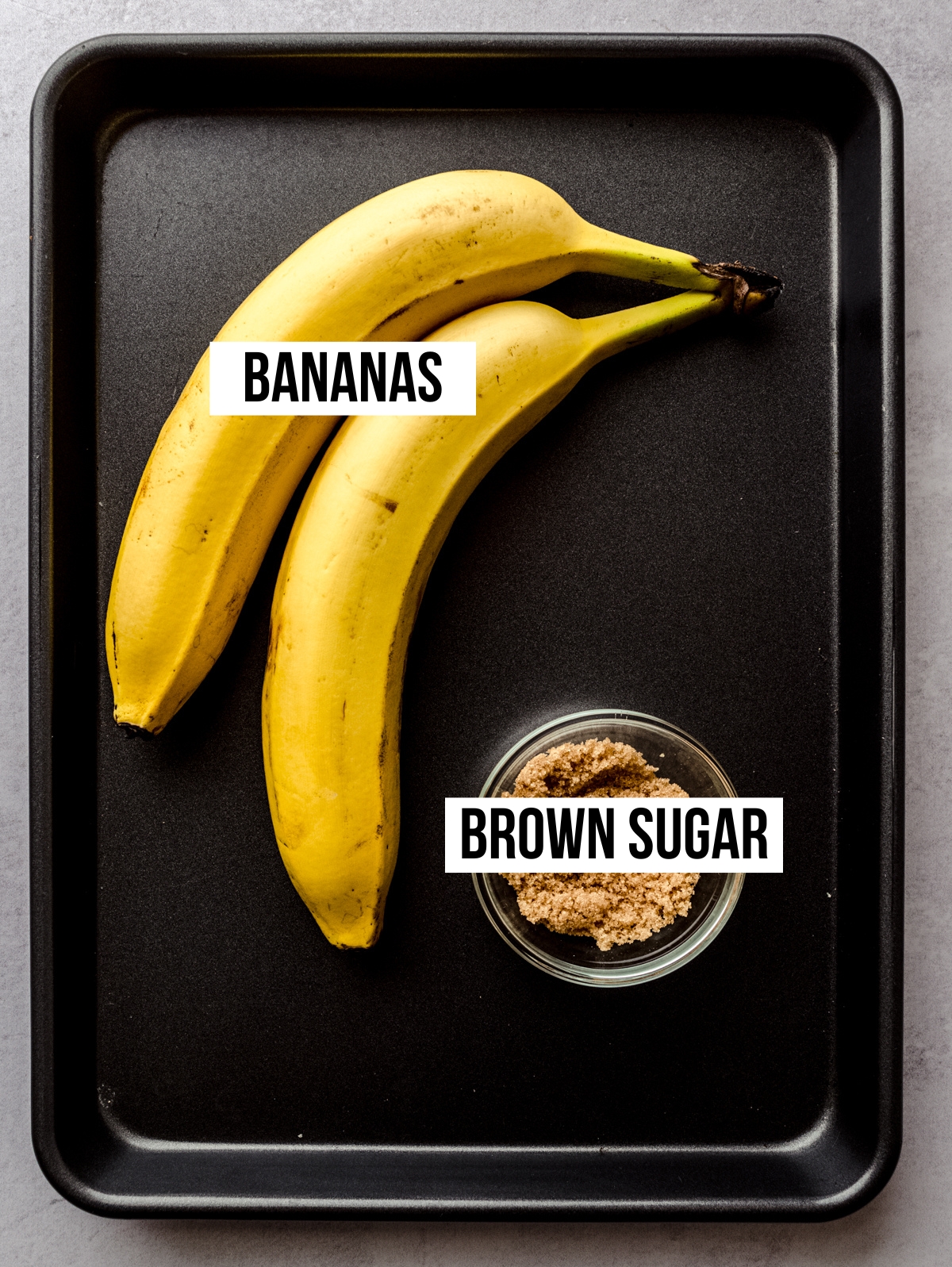 Aerial photo of ingredients to make roasted bananas on a dark baking sheet with text overlay labeling each ingredient.