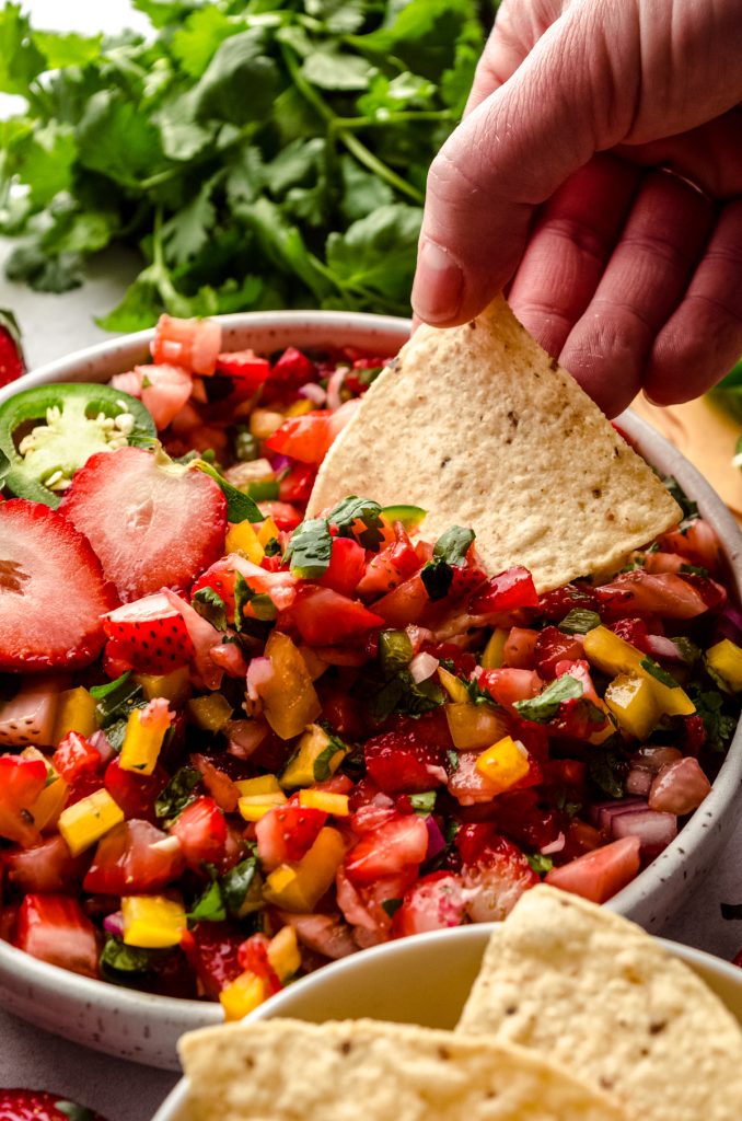 Someone is dipping a tortilla chip into a bowl of fresh strawberry salsa.