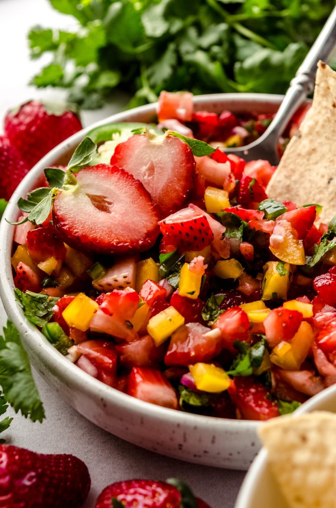 A bowl of fresh strawberry salsa with fresh strawberries and chips around it and a chip dipped into a bowl.
