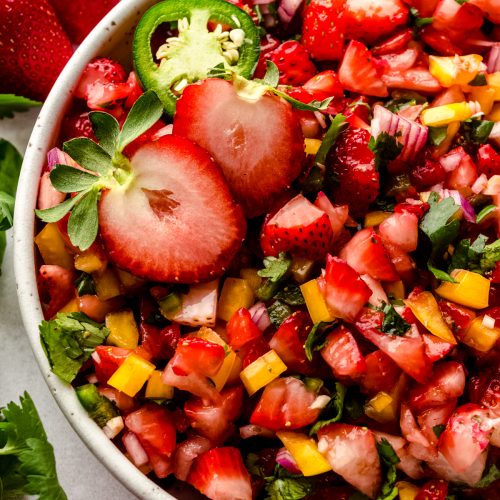 A bowl of fresh strawberry salsa with fresh strawberries and chips around it.