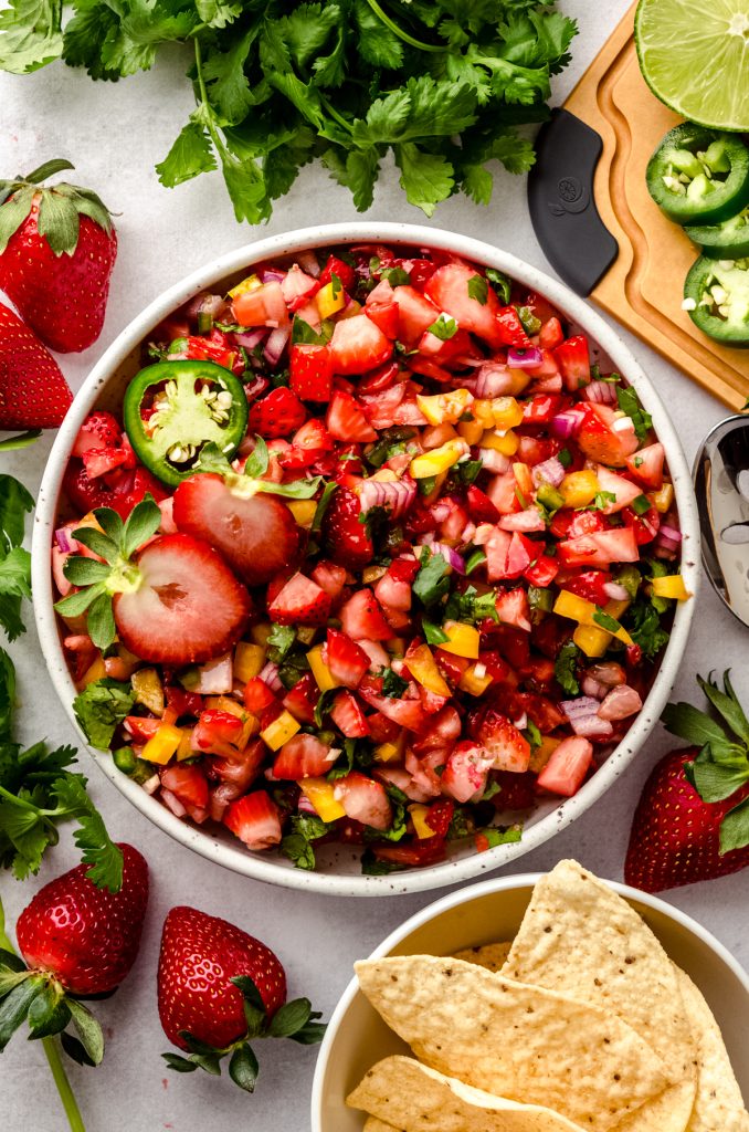 An aerial photo of a bowl of fresh strawberry salsa with fresh strawberries and chips around it.