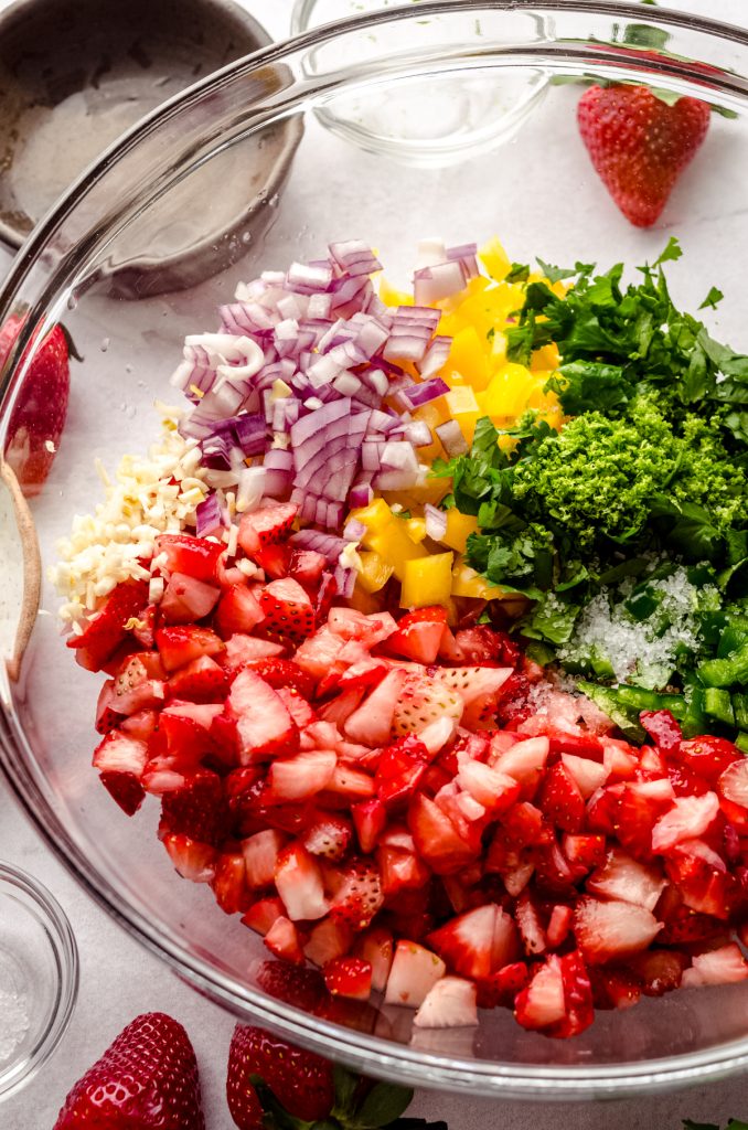 Strawberries, red onion, yellow bell pepper, lime, jalapeño, garlic, and cilantro in a bowl to make strawberry salsa.