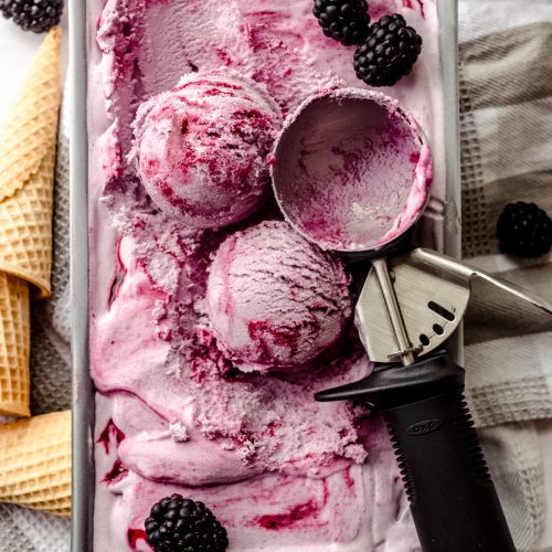 Aerial photo of blackberry ice cream in a container and scoops have been formed with an ice cream scoop and there are cones lying around the container.