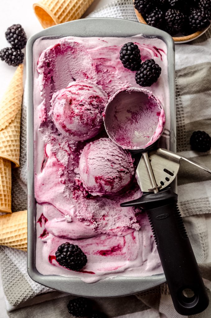 Aerial photo of blackberry ice cream in a container and scoops have been formed with an ice cream scoop and there are cones lying around the container.