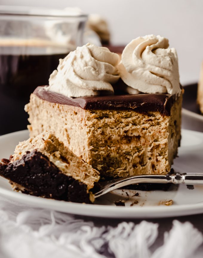 A slice of coffee cheesecake on a plate with a fork and a bite has been taken out of it.