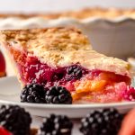 A slice of blackberry peach pie on a plate with blackberries on the side of the plate and the full pie in the background and peaches and blackberries around it.