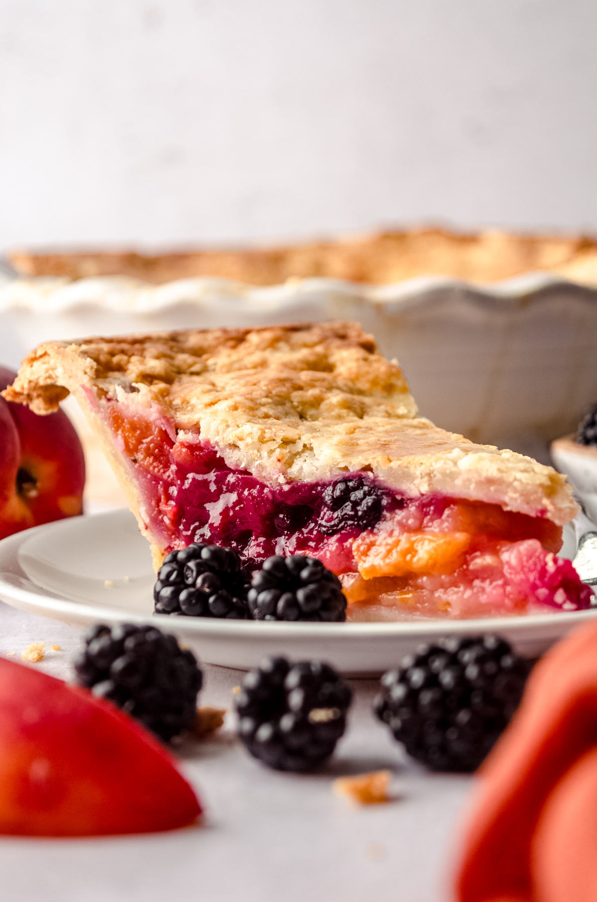 A slice of blackberry peach pie on a plate with blackberries on the side of the plate and the full pie in the background and peaches and blackberries around it.