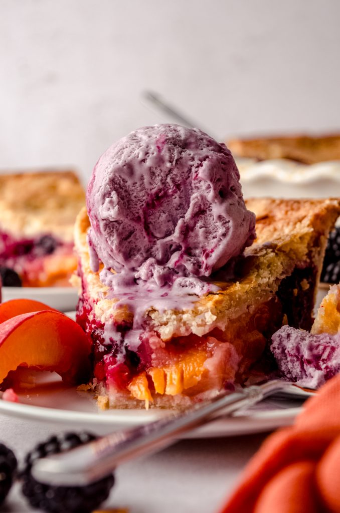 A slice of blackberry peach pie on a plate with a bite taken out of it and the full pie in the background and peaches and blackberries around it and a scoop of blackberry ice cream is sitting on top and starting to melt.