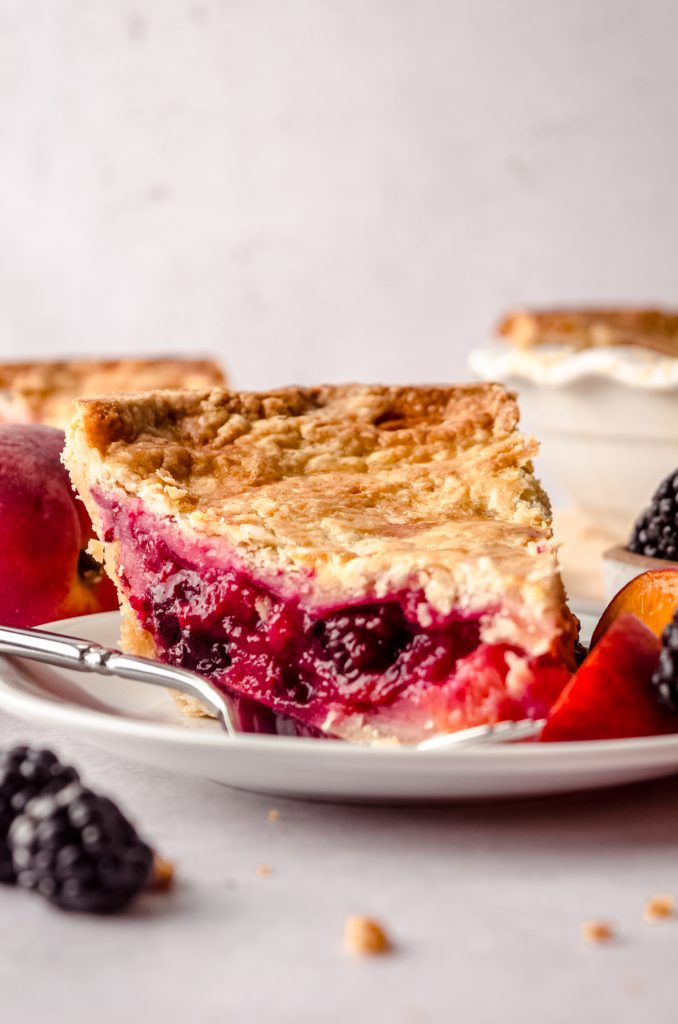 A slice of blackberry peach pie on a plate with blackberries on the side of the plate and the full pie in the background and peaches and blackberries around it.