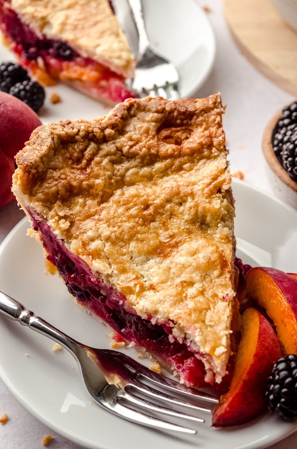 A slice of blackberry peach pie on a plate with blackberries on the side of the plate and the full pie in the background and peaches and blackberries around it.