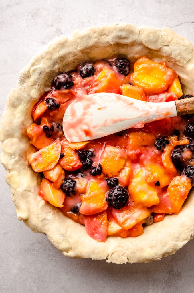 Aerial photo of the filling for blackberry peach pie in the pie crust with a spatula spreading the filling around.