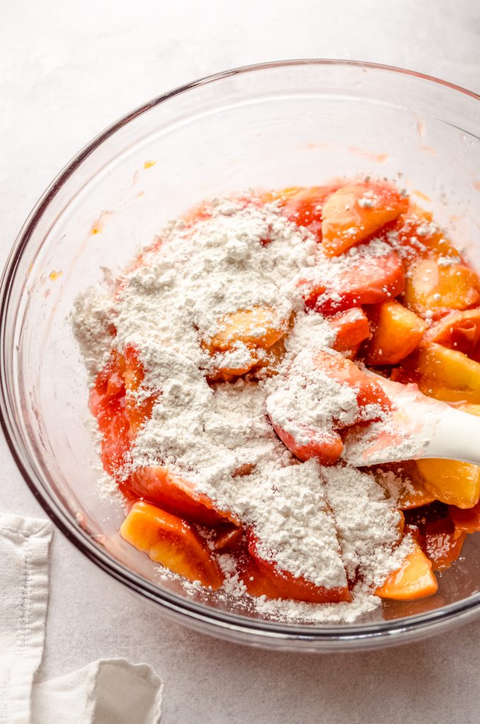 Peaches in a bowl with flour on them and a spatula ready to make peach blackberry pie.