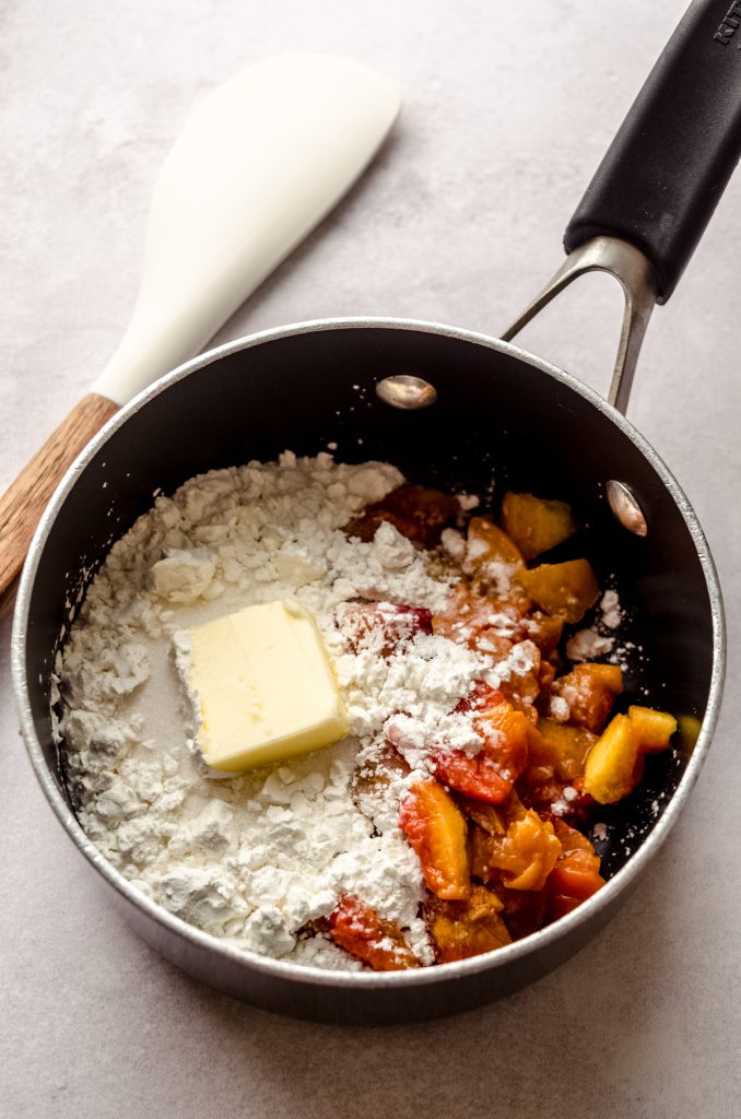 A saucepan with peaches, cornstarch, butter, and a spatula ready to cook to make peach blackberry pie.