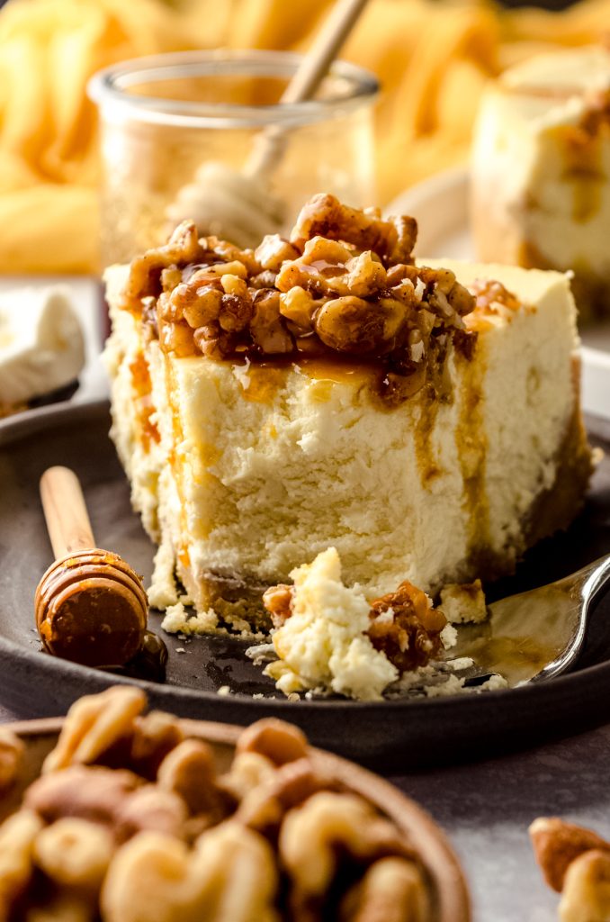 A slice of goat cheese cheesecake sitting on a plate with a honey stick and a fork and a bite has been taken out of the slice.