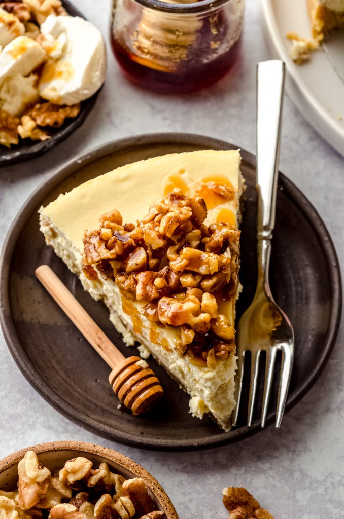 A slice of goat cheese cheesecake sitting on a plate with a honey stick and a fork and the slice has a honey walnut topping that is dripping down the side of the slice.