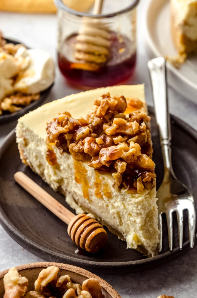 A slice of goat cheese cheesecake sitting on a plate with a honey stick and a fork and the slice has a honey walnut topping that is dripping down the side of the slice.