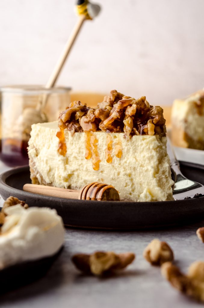 A slice of goat cheese cheesecake sitting on a plate with a honey stick and the slice has a honey walnut topping that is dripping down the side of the slice.