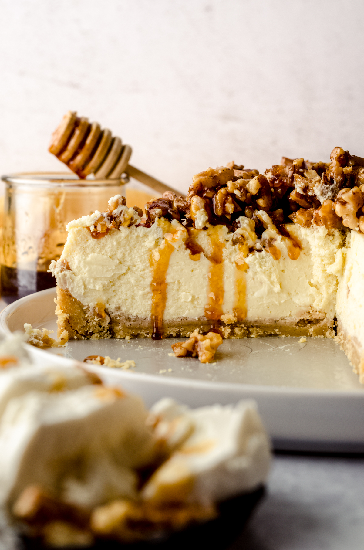 A sliced goat cheese cheesecake sitting on a plate with a jar of honey and a honey stick in the background and a plate of goat cheese in the foreground.