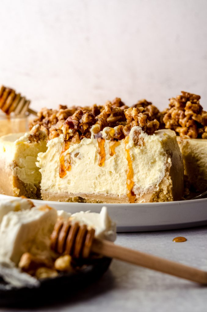 A slice of goat cheese cheesecake sitting on a platter and the slice has a honey walnut topping that is dripping down the side of the slice.