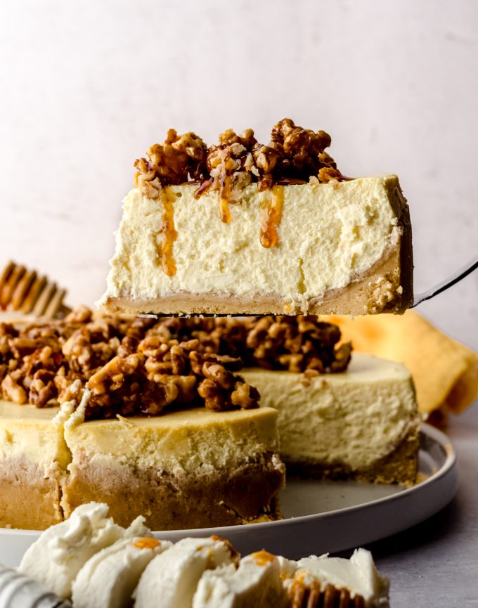 Someone is using a server to lift a slice of goat cheese cheesecake and the slice has a honey walnut topping that is dripping down the side of the slice.