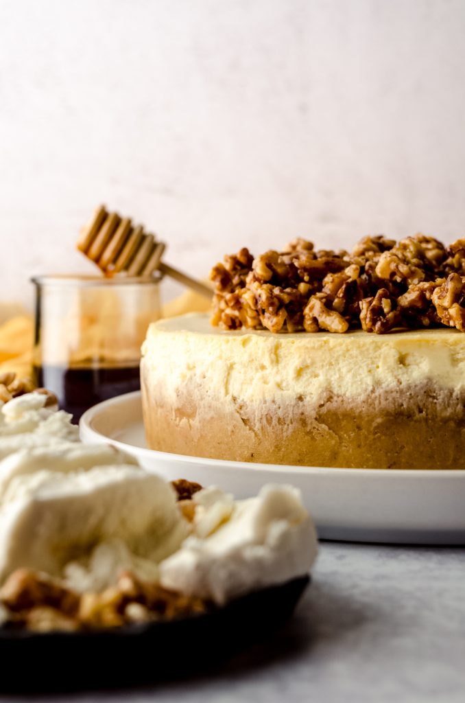 A goat cheese cheesecake sitting on a plate with a jar of honey and a honey stick in the background and a plate of goat cheese in the foreground.