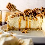 A sliced goat cheese cheesecake sitting on a plate with a jar of honey and a honey stick in the background and a plate of goat cheese in the foreground.