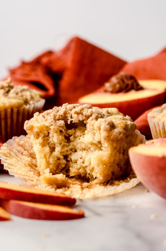 A peach muffin on a surface with an orange kitchen towel in the background with peach slices around it and a bite has been taken out of the muffin.