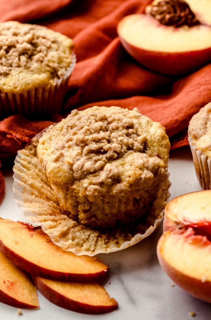 A peach muffin on a surface with an orange kitchen towel in the background with peach slices around it.